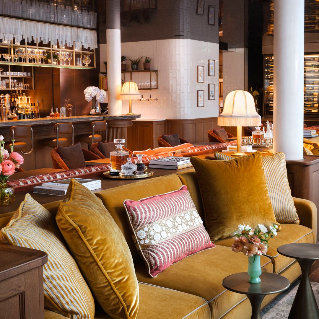 Eine gemütliche Couch mit Zierkissen, eine farbenfrohe Blumenvase auf dem Tisch, stilvolle Tischlampen, gerahmte Kunstwerke und die Neue Hoheit Bar im Hintergrund.